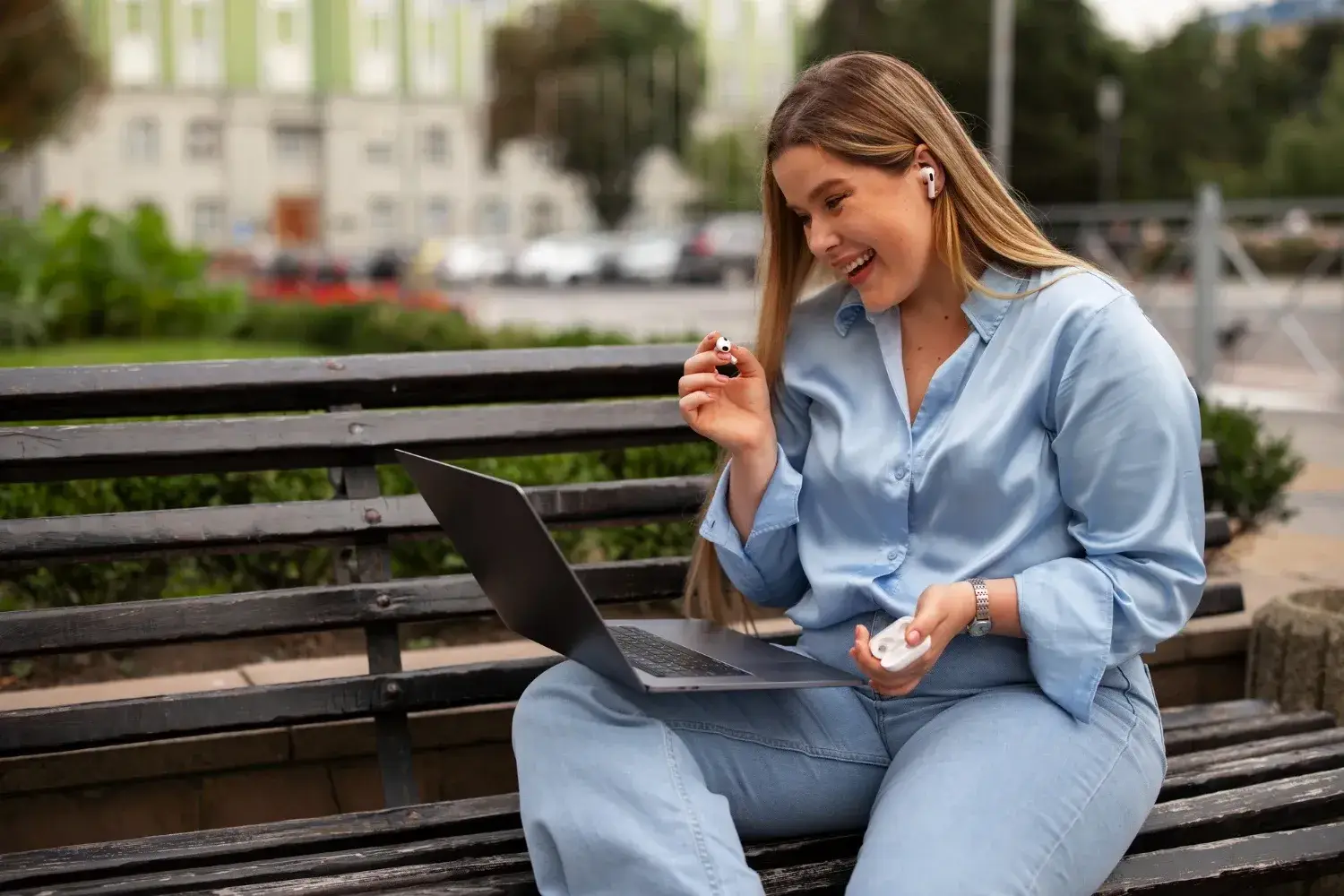 A woman sitting on a bench using a laptop while holding wireless earbuds in her hand.