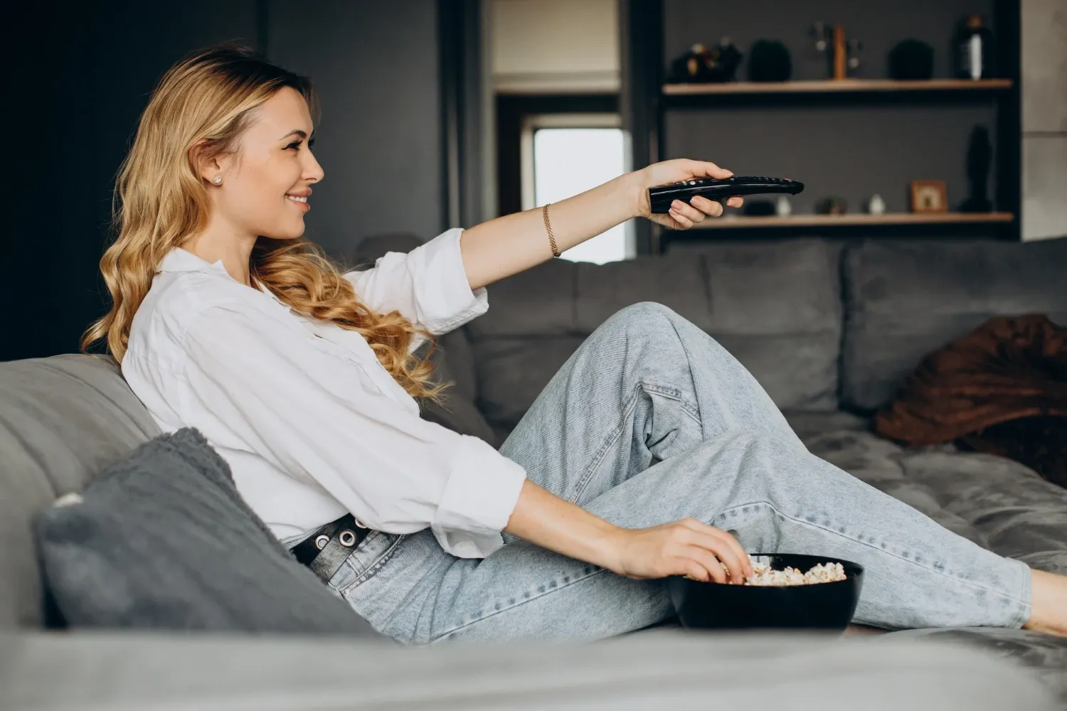 Woman sitting on a couch holding a remote control and a bowl of popcorn while watching TV.